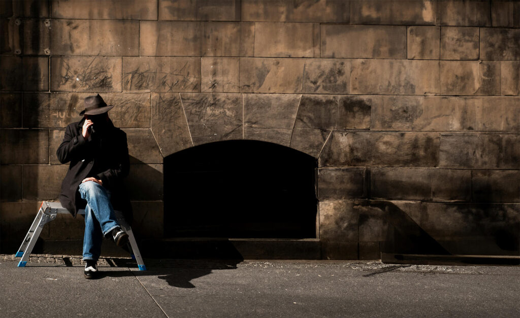 Une personne assise sur une échelle pliante, partiellement ombragée, portant un manteau sombre et un chapeau fedora, parlant au téléphone portable devant un mur de briques texturé, éclairé par la lumière du soleil urbain.